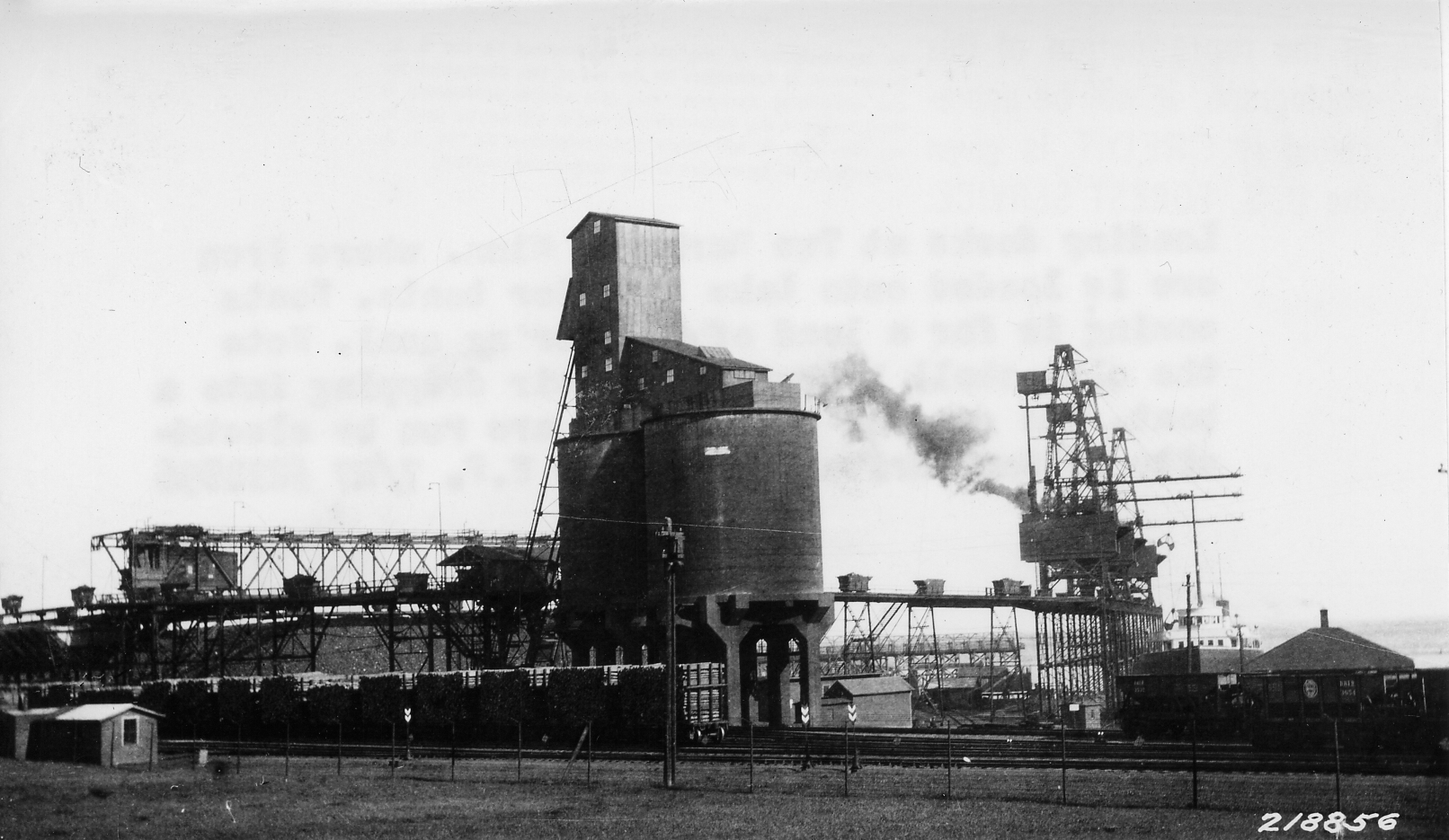 Scope and content: Original caption: Loading docks at Two Harbors, Minn where iron ore is loaded onto Lake Superior boats. Boats coming in for a load of iron bring coal. Note the clam-shell bucket in the air dropping into a boat. The cars on the trestle are run by electricity without drivers.General notes: The original caption is incorrect. This is the Duluth and Iron Range Railroad coal facility at Agate Bay in Two Harbors Minnesota. The boat is having coal unloaded. The tram cars are on a continuous cable drive system that loops them around to the bridge crane that covers the storage field. The storage field could hold 150,000 tons of mine run coal. The concrete towers are a crushing and holding facility. The large locomotives had worm gear coal feeders and could not handle mine run coal. Each tower could hold 10,000 tons of crushed coal. I am sure the Forest Service has interest in this photograph because of the volume of pulpwood logs on flat cars next to the coal towers.(Mel Sando Lake County Historical Society)